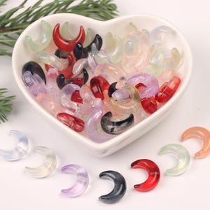 May include: A heart-shaped bowl filled with a variety of colorful crescent moon-shaped beads. The beads are made of translucent glass and come in shades of red, pink, purple, green, and black.