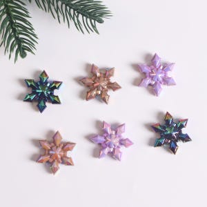 Könnte beinhalten: Sechs schimmernde Schneeflocken-Ornamente in verschiedenen Farben, darunter Lila, Blau und Braun. Die Schneeflocken sind auf einem weißen Hintergrund angeordnet.