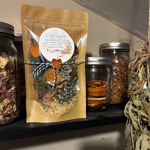 May include: A brown kraft paper pouch containing a Sweater Weather Simmer Pot mix, tied with green and white twine. The label features an apple icon and instructions to add contents to a pot with 3 cups of water and simmer on low, adding water as needed.