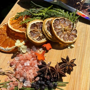 May include: A collection of dried orange slices, star anise, pink Himalayan salt crystals, dark juniper berries, and green cedar sprigs. These botanical ingredients are arranged together, with a holographic pouch visible at the top edge.