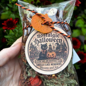 May include: A clear plastic bag filled with dried botanical potpourri, tied with orange and black twine. A round beige tag features a black cat, a pumpkin, and a haunted house illustration with the text Nostalgic Halloween, Smells Like Memories.