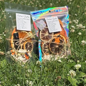 May include: Two clear plastic pouches containing dried fruit slices, including orange and lime, and botanical ingredients. Each pouch features a white label with the text Tropical Summer Break Simmer Pot and is tied with twine. One pouch has a holographic finish.