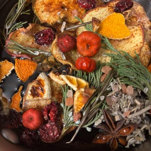 May include: A simmering potpourri mixture containing dried apple slices, orange peels, whole star anise, sprigs of green rosemary, cedar needles, small red berries, and pink salt crystals submerged in a dark liquid base within a metal container.