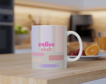 May include: White ceramic coffee mug with the text "coffee club" in pink. The mug features a pastel color block design with vertical stripes and rectangular shapes in shades of pink, lavender, and beige. The mug is on a wooden surface.