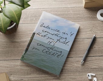 May include: A notebook with a serene ocean scene and the handwritten quote, "Balance isn't something that you find. It's something you CREATE!" A pen, earbuds, and a plant are on a wooden surface.