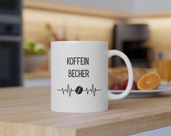 May include: White ceramic coffee mug with black text that reads "Koffein Becher" and a stylized heartbeat with a coffee bean in the center.
