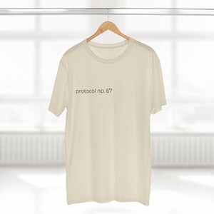 Puede incluir: Camiseta de color crema colgada de una percha de madera. La camiseta tiene mangas cortas y cuello redondo. El texto "protocol no. 67" está impreso en color oscuro en la parte delantera.
