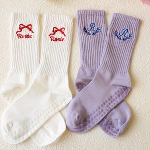 May include: Two pairs of socks. One pair is white with red embroidered bows and the name "Rosie". The other pair is lavender with blue embroidered "R" initials and laurel wreaths. Both pairs have grip dots on the soles.