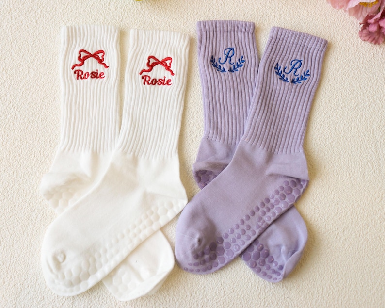 May include: Two pairs of socks. One pair is white with red embroidered bows and the name "Rosie". The other pair is lavender with blue embroidered "R" initials and laurel wreaths. Both pairs have grip dots on the soles.