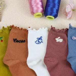 Può includere: Cinque paia di calzini colorati con dettagli ricamati. I calzini sono in tonalità di verde, marrone, bianco, malva e blu. Ogni calzino ha un design unico, tra cui ciliegie, la parola "Cocoa", cuori, un coniglio e il nome "Gemma".
