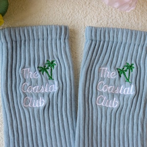 May include: A pair of light blue ribbed socks. Each sock features the embroidered text "The Coastal Club" in white, with a green palm tree graphic above the text. The socks are made of a soft, ribbed material.