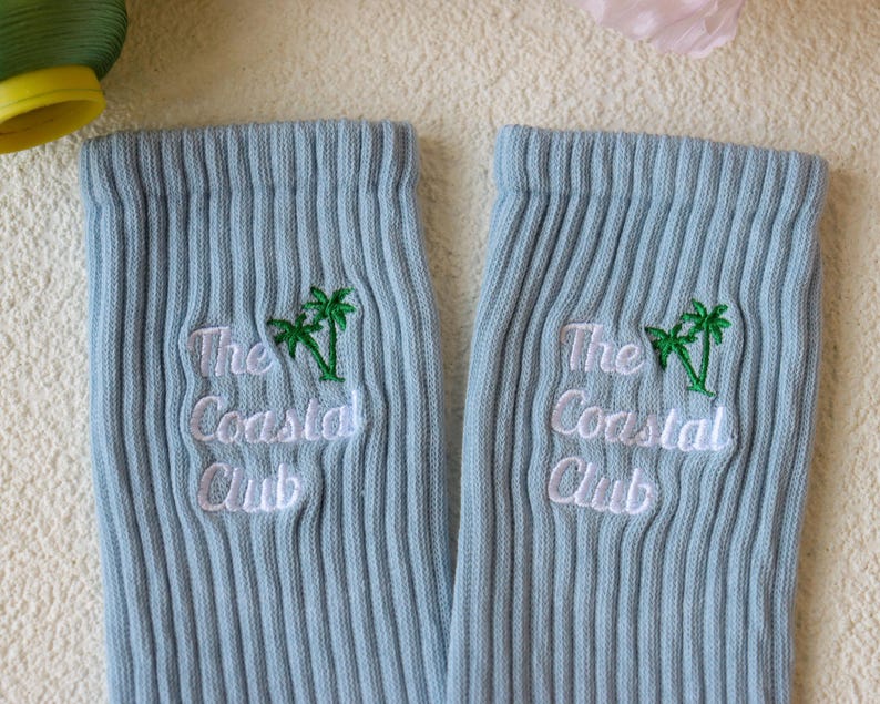 May include: A pair of light blue ribbed socks. Each sock features the embroidered text "The Coastal Club" in white, with a green palm tree graphic above the text. The socks are made of a soft, ribbed material.