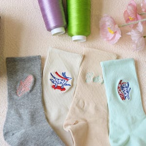 May include: A selection of patterned socks in various colours. The socks feature embroidered designs, including a corn, a dog on a skateboard, and a cat. The socks are in shades of grey, cream, and light green. Spools of thread and flowers are in the background.