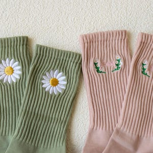 May include: Two pairs of ribbed socks. One pair is light green with embroidered white daisies with yellow centers. The other pair is pink with embroidered text and floral accents. The socks are made of a soft, comfortable material.