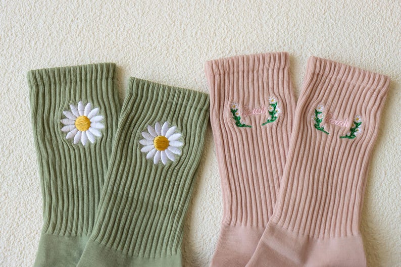 May include: Two pairs of ribbed socks. One pair is light green with embroidered white daisies with yellow centers. The other pair is pink with embroidered text and floral accents. The socks are made of a soft, comfortable material.