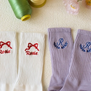 May include: White ribbed socks embroidered with a red bow and the name "Rosie". Two lavender ribbed socks are embroidered with the letter "R" and a blue laurel wreath. Sewing thread and flowers are in the background.