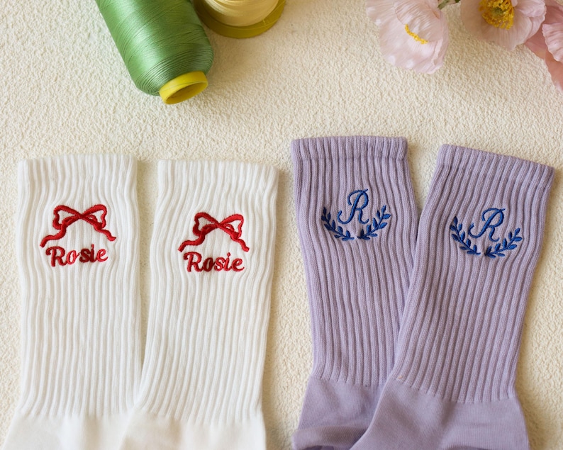 May include: White ribbed socks embroidered with a red bow and the name "Rosie". Two lavender ribbed socks are embroidered with the letter "R" and a blue laurel wreath. Sewing thread and flowers are in the background.