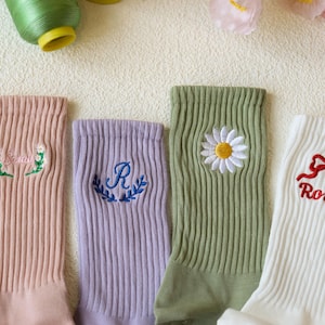 May include: Four pairs of ribbed socks in pastel colors, each with embroidered designs. One pair is peach with floral embroidery, another is lavender with a letter "R", a third is green with a daisy, and the last is white with a red bow and the name "Rosie".