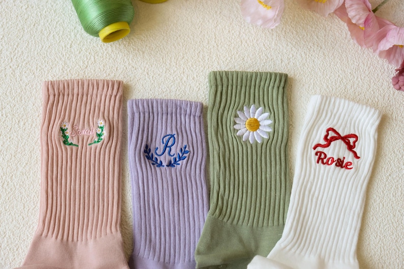 May include: Four pairs of ribbed socks in pastel colors, each with embroidered designs. One pair is peach with floral embroidery, another is lavender with a letter "R", a third is green with a daisy, and the last is white with a red bow and the name "Rosie".
