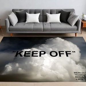Può includere: Un tappeto bianco e nero con un design a nuvole e il testo "KEEP OFF" stampato su di esso. Il tappeto si trova in un soggiorno con un divano grigio e un tavolino in legno.