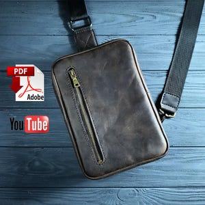 May include: A dark brown leather crossbody bag featuring a vertical zippered pocket with a brass-toned metal zipper pull. The bag includes a wide black adjustable fabric strap attached with metal hardware. The design is rectangular with rounded corners.