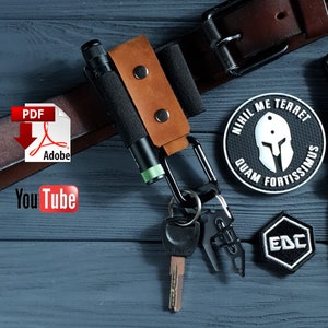May include: A brown leather belt loop with a black flashlight and a black carabiner holding keys. The belt loop has a brown leather strap with two silver rivets. A black and green flashlight is secured in a black fabric loop.