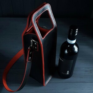 May include: Black leather wine tote with red trim and handle. The tote has a rectangular shape with a handle at the top and a shoulder strap. A bottle of red wine sits next to the tote.