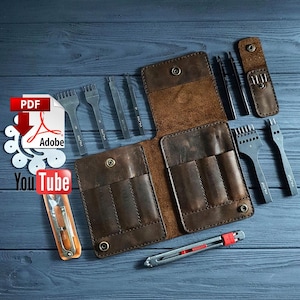 Op de afbeelding: Een bruine leren portemonnee, open om de vakken te laten zien, omringd door leerbewerkingstools. Deze omvatten ponsen en een hobbymes. De gereedschappen zijn van metaal met houten handvatten. De afbeelding is geplaatst tegen een blauwe houten achtergrond.