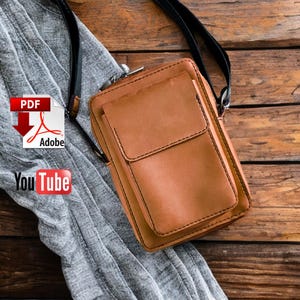 Può includere: Borsa a tracolla in pelle marrone con cinturino nero. La borsa ha una forma rettangolare con una tasca frontale e una chiusura a zip. La borsa è su una superficie di legno.