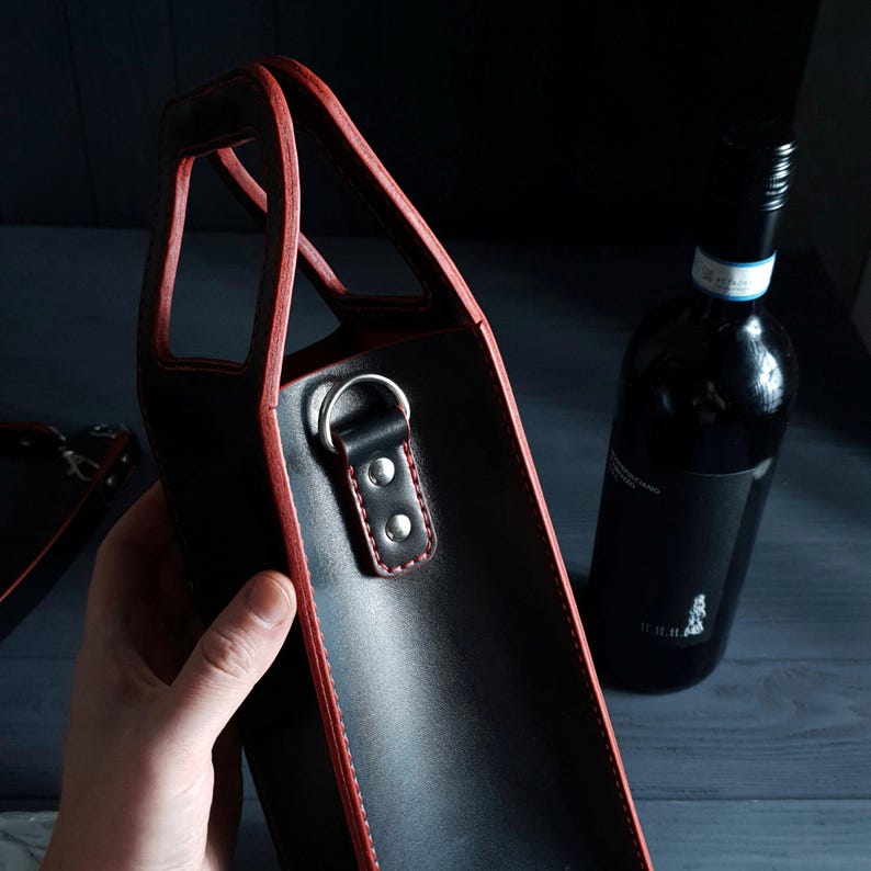 May include: Black leather wine bottle carrier with red trim and a handle. The carrier has a silver metal ring and a small leather strap with two silver rivets. A dark wine bottle is in the background.