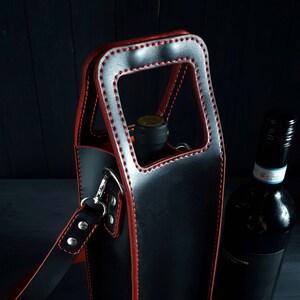May include: Black leather wine bottle carrier with red stitching and a matching shoulder strap. The carrier has a rectangular handle and holds a wine bottle. A second wine bottle is next to the carrier.