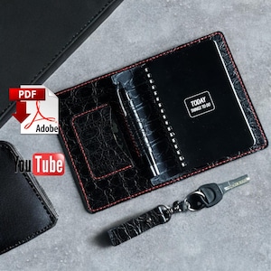 以下が含まれることがあります： 赤いステッチが施された黒革のノート、ペン、キーホルダー。 ノートには「TODAY THINGS TO DO」の文字が表紙に書かれています。 ノートは開いており、スパイラル綴じのメモ帳が現れています。 キーホルダーは鍵に付いています。