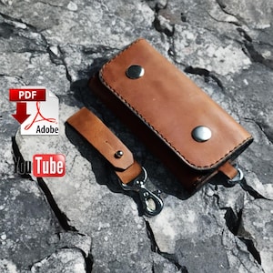 May include: A brown leather key holder and key fob set with silver-tone metal hardware. The key holder is rectangular with two snap closures. The key fob has a snap closure and a metal clasp. The set is on a grey stone surface.