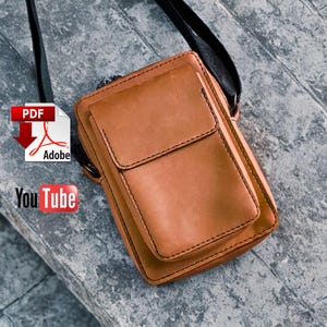 Könnte beinhalten: Hellbraune Leder-Umhängetasche mit Klappe und schwarzen Nähten. Die Tasche hat einen langen schwarzen Riemen und eine kleine Vordertasche. PDF- und YouTube-Symbole im Bild.