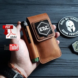 Puede incluir: Un estuche de cuero marrón para bolígrafo EDC (Every Day Carry) con un parche EDC en blanco y negro. El estuche contiene un bolígrafo negro con un acento verde. También es visible un parche en blanco y negro con el texto "NIHIL ME TERRET QUAM FORTISSIM".