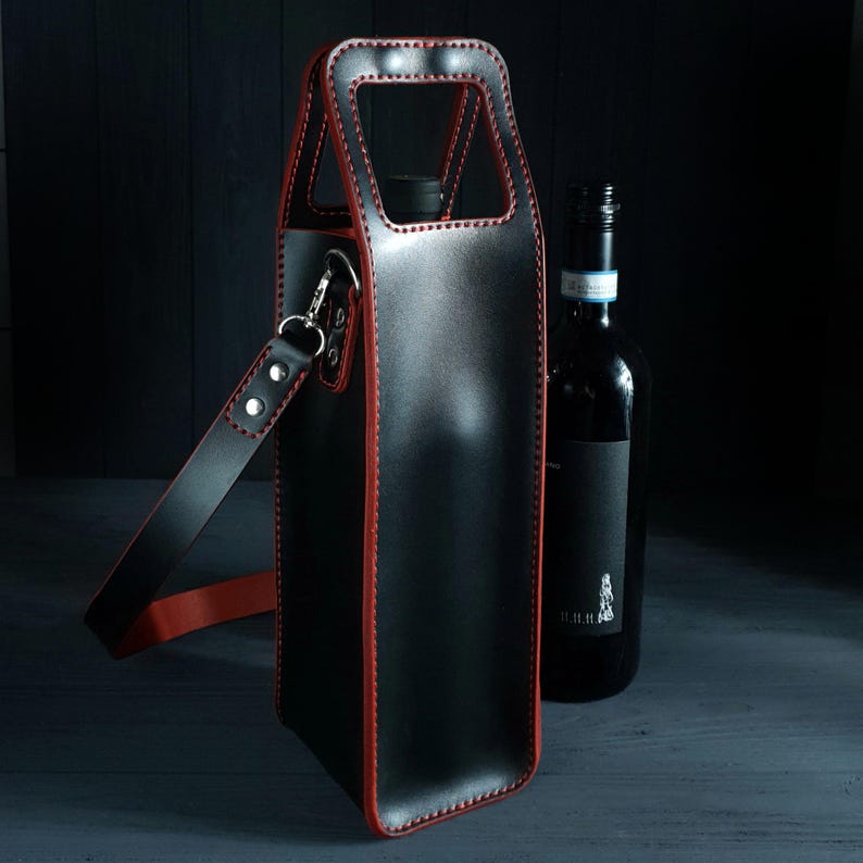 May include: Black leather wine bottle carrier with red stitching and a shoulder strap. The carrier has a handle at the top and a silver clasp. A dark wine bottle is next to the carrier.