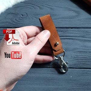 May include: A tan leather keyring with a silver clasp and rivet. The keyring is held in a hand against a dark grey background. The image also includes a PDF Adobe icon and a YouTube logo.
