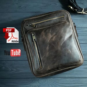 Puede incluir: Un bolso de cuero marrón oscuro con silueta rectangular, un bolsillo horizontal con cremallera en la parte superior y un bolsillo vertical con cremallera en el panel frontal. El bolso incluye una correa negra extraíble con herrajes metálicos.