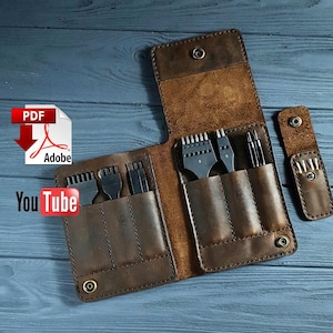Könnte beinhalten: Ein brauner Leder-Werkzeugorganizer, offen, um verschiedene Metallwerkzeuge zu zeigen. Der Organizer hat mehrere Taschen und wird mit Druckknöpfen befestigt. Die Werkzeuge umfassen Punzen und andere Lederverarbeitungswerkzeuge. Der Hintergrund ist eine blaue Holzoberfläche.