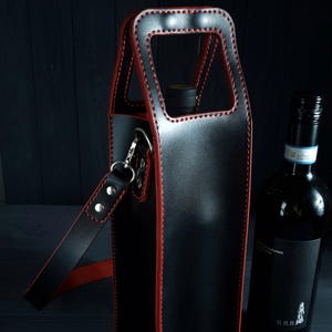 May include: Black leather wine bottle carrier with red stitching and a shoulder strap. The carrier has a handle and a silver-colored clasp. A dark wine bottle is next to the carrier.