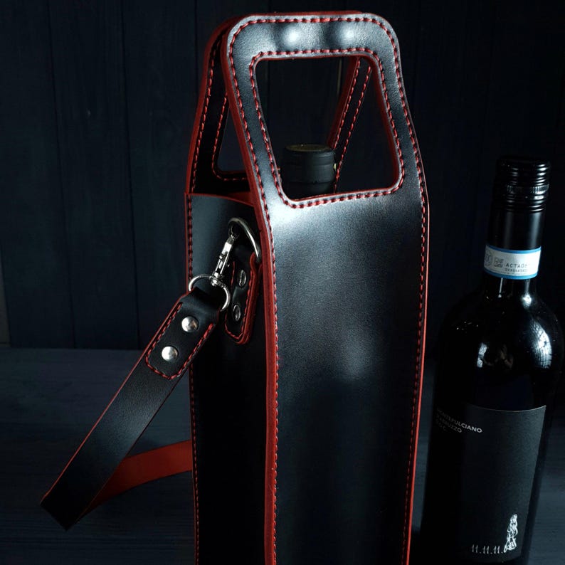 May include: Black leather wine bottle carrier with red stitching and a shoulder strap. The carrier has a handle and a silver-colored clasp. A dark wine bottle is next to the carrier.