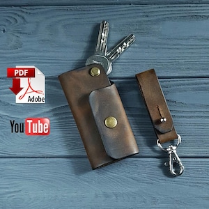 May include: A brown leather key holder with a snap closure and a matching key fob with a silver clasp. Two keys are inserted into the holder. The key holder is on a blue wooden surface.