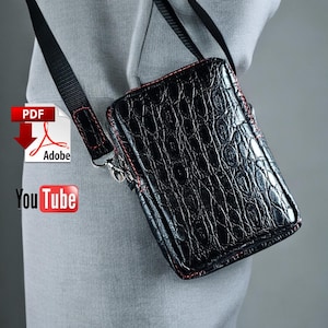 May include: A black leather crossbody bag with a red stitch detail and a crocodile-embossed pattern. The bag has a zipper closure and a long adjustable strap.