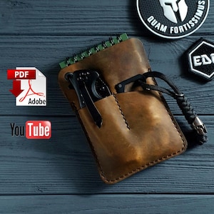 Op de afbeelding: Een bruine leren notitieboekhoes met een vak, met een klein notitieboekje en twee zwarte vouwmesjes. Een zwart koord met een schedelbedel is bevestigd. De achtergrond is een donkerblauwe houten ondergrond.