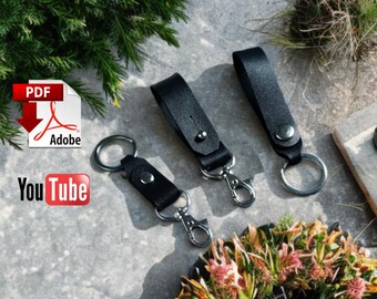 May include: Three black leather keychains with silver-colored hardware are displayed on a textured surface. Each keychain features a loop and a clasp. The keychains are arranged near greenery and a floral arrangement.