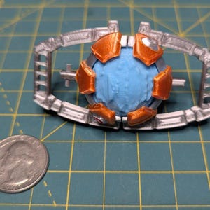 May include: A detailed, 3D-printed model featuring a central blue sphere encased in orange and silver components. The model is set against a green grid background, with a U.S. quarter coin for size comparison.