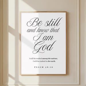 Może przedstawiać: Oprawiony wydruk z napisem "Be still and know that I am God" w eleganckiej czcionce. Wydruk zawiera również tekst "I will be exalted among the nations, I will be exalted in the earth. PSALM 46:10". Rama jest w kolorze jasnobrązowym.