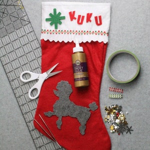 May include: A red Christmas stocking with the name "KUKU" in red letters. The stocking is decorated with a green felt snowflake and a gray poodle applique. Craft supplies include scissors, glue, tape, and sequins.