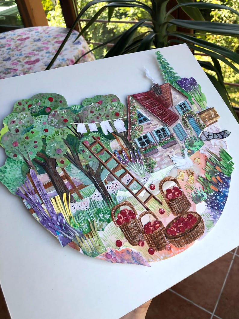 May include: A layered paper collage artwork depicting a rural scene with a cottage, apple trees, and a wooden ladder. The image features three woven baskets filled with red apples, a white goose with chicks, laundry on a line, and green foliage.