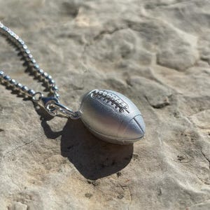 Könnte beinhalten: Ein silberner Football-Anhänger hängt an einer feinen Kugelkette. Der Anhänger ist eine detaillierte Miniatur eines Footballs, komplett mit Schnürsenkeln. Die Halskette und der Anhänger werden auf einer strukturierten, hellfarbigen Steinoberfläche präsentiert.